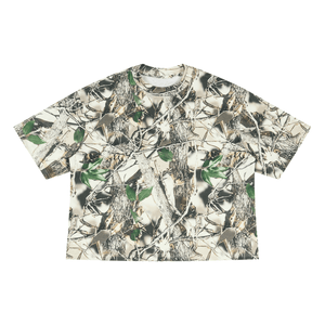 A short-sleeve crewneck t-shirt featuring a realistic woodland camouflage pattern with tree bark, branches, leaves, and grass in shades of beige, brown, green, and gray, laid flat against a white background.