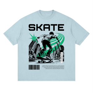 A light blue crewneck t-shirt featuring a large graphic print on the front. The graphic displays the word 'SKATE' in bold black letters above an illustration of a skateboarder in black and green, with a cityscape background, a green starburst, and a barcode with text below.