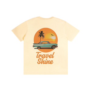 A light cream-colored short-sleeve t-shirt featuring a retro graphic of a teal classic car driving in front of an orange sunset with a palm tree, and the text 'Travel Shine' printed below.