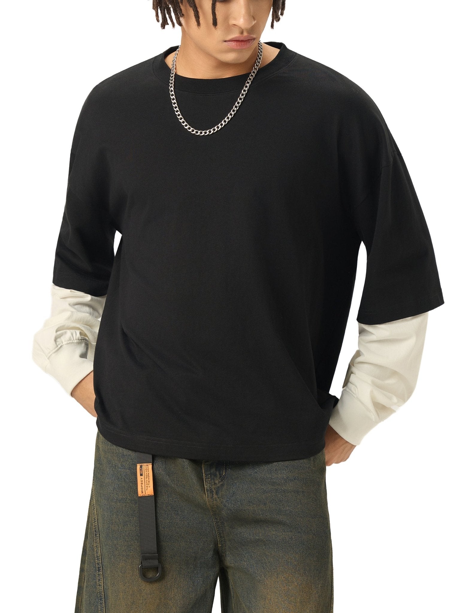 Person wearing a black long-sleeve shirt over a white shirt with visible tattoos, paired with dark pants on a white background.