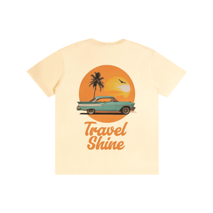 A light cream-colored short-sleeve t-shirt featuring a retro graphic of a teal classic car driving in front of an orange sunset with a palm tree, and the text 'Travel Shine' printed below.