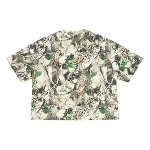 A short-sleeve button-up shirt featuring an all-over woodland camouflage pattern with branches, leaves, and bark in shades of beige, brown, green, and gray, shown flat against a plain white background.